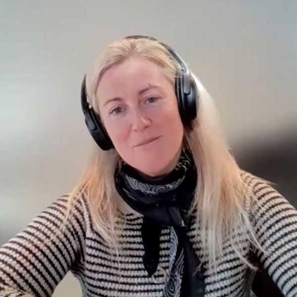 En blond kvinna med hörlurar, randig tröja och en knuten sjal runt halsen. Bakgrunden är suddig.