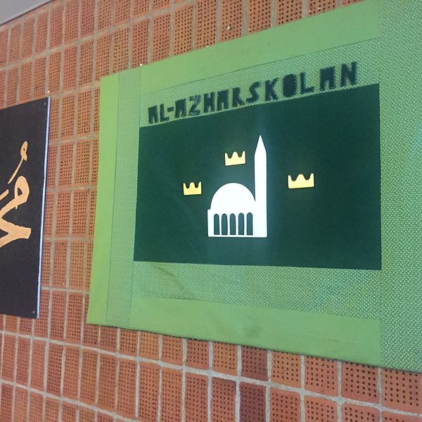 Två tygtavlor på tegelvägg. En svart med arabisk text på och en grön där det står Al-Azharskolan.