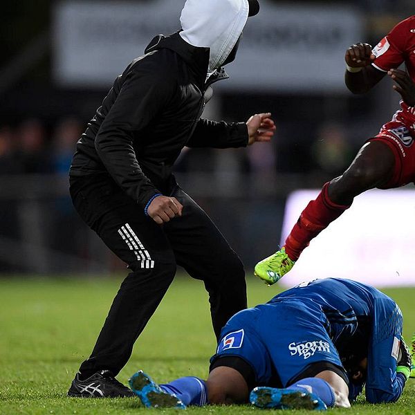 Maskerad man på en fotbollsplan. En spelare ligger på marken, en annan är på väg mot den maskerade mannen.