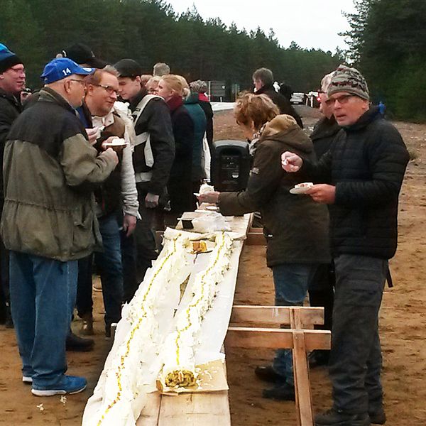 En lång rulltårta ligger på ett avlångt provisoriskt bord. Människor äter tårta. De är utomhus. Höga granar syns i bakgrunden.