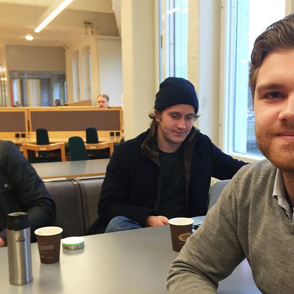 Tre studenter sitter vid ett bord. En ung man tittar in i kameran, han är till höger i bild.