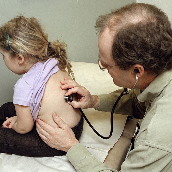En barnläkare undersöker en flicka i 3-års ålder på ett undersökningbord. Flickan sitter upp.