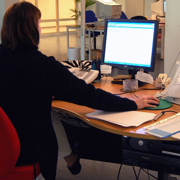 Kvinna sitter på en röd kontorsstol vid en arbetsplats och tittar mot en datorskärm, har högra handen på datormusen. På skrivbordet ligger papper och penna samt en telefon och hörlurar.