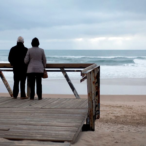 Äldre par som står på altan och blickar ut mot sandstrand och havet. Ska symbolisera pensionärer som bosatt sig i  Portugal som snart kan komma att behöva betala skatt i landet.