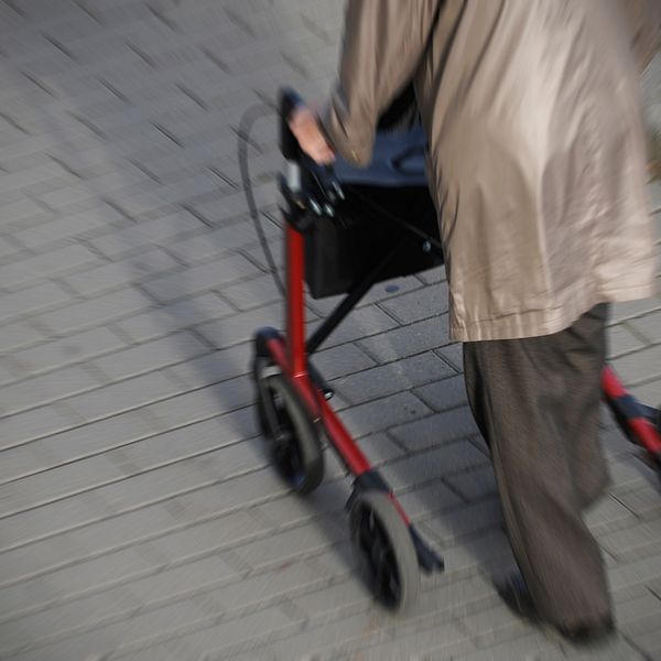 äldre person med brun kappa går med röd rollator