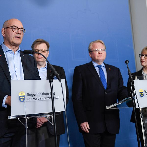 En man med glasögon bakom ett talarpodium pratar på en presskonferens, två män och en kvinna står lite bakom honom.