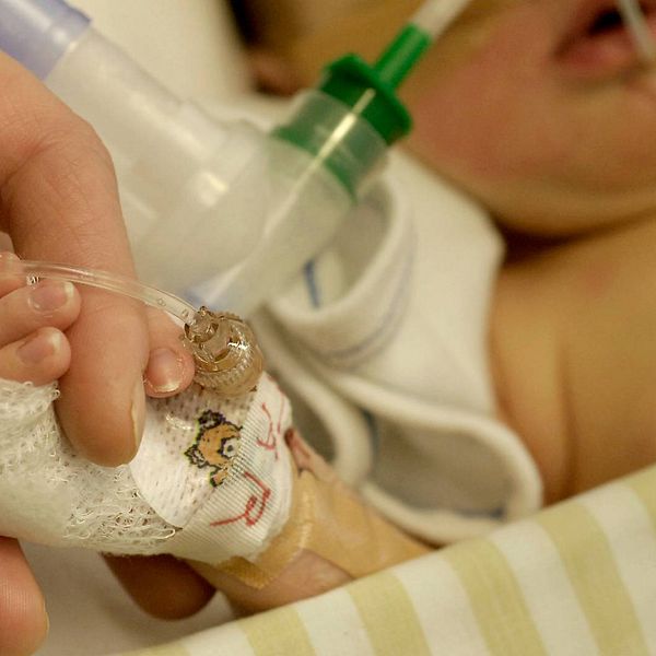 En vuxen hand håller i ett litet barns bandagerade hand på sjukhus. Barnet har slangar i näsan.