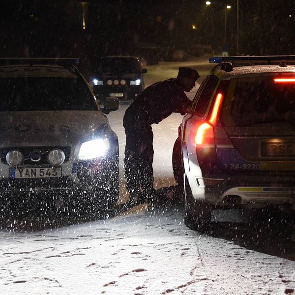 Två polisbilar står på en väg i mörker och snö, en polisman pratar med föraren i ena bilen.