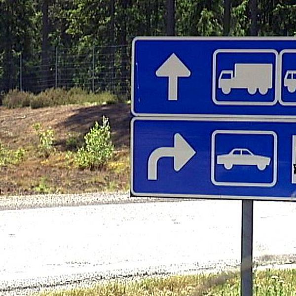 Skog i bakgrunden med blå vägskylt vid avfart till en rastplats. På den blåa vägskylten syns olika fordonskyltar för lastbil, bussar och personbilar.