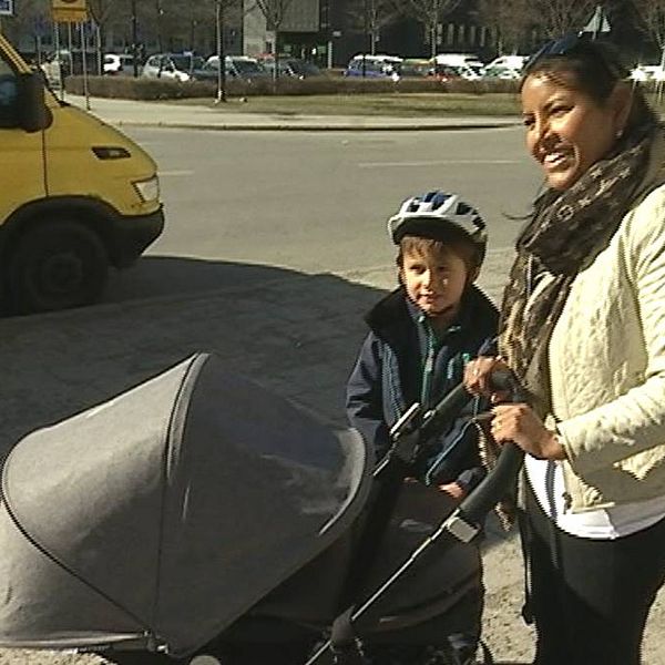 En kvinna står på trottoar med en barnvagn och ett litet barn med cykelhjälm.