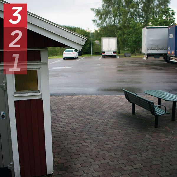 En parkeringsplats vid en rastplats med flera lastbilar parkerade. Till vänster i bilden finns en röd byggnad med en dörr märkt ”Toalett” för handikappanpassning. I förgrunden syns ett par bänkar och ett bord på en stenbelagd yta.
