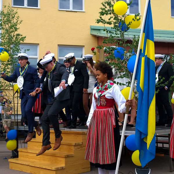 Ett gäng studenter står samlande med studentmössor på. Ballonger och Sverigeflaggor pryder scenen de står på.