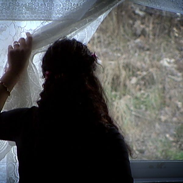 En person med långt hår står bortvänd mot kameran och tittar ut genom ett fönster med spetsgardiner.