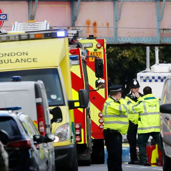 Tre polismän i gula jackor står och pratar med varandra intill en brandbil, en ambulans och en polisbil intill en bro vid en tunnelbaneskylt i London.