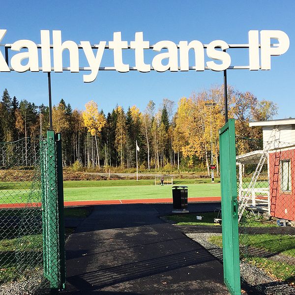 En grön grind står öppen, där syns träd och en gräsplan. En stor skylt i mitten av bilden ”Kalhyttans IP”.