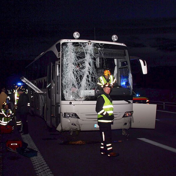 Vit buss med stora skador på framrutan. Räddningspersonal. Människor håller om varandra vid sidan av motorvägen.