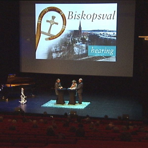 På en scen står tre personer under en skylt där det står Biskopsval hearing