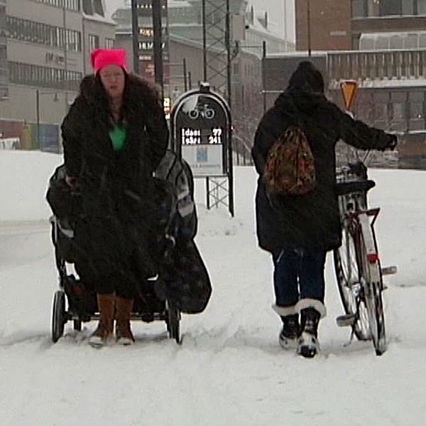 Två personer i svarta jackor går längs en väg där snön yr omkring dem.