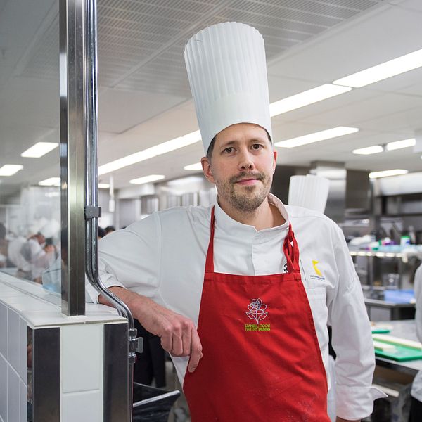 Daniel Roos står i ett kök med massa kockar som arbetar bakom. Han har på sig vit rockmössa, vit kockskjorta och rött förkläde.