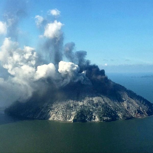 Bildtext vulkanen Kadovar, omgiven av vatten, med blå himmel bakom. Från toppen kommer kraftig rök.