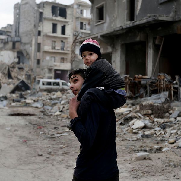 En ung man med ett barn sittandes på axlarna ser sig omkring i en sönderbombad del av syriska staden Aleppo.