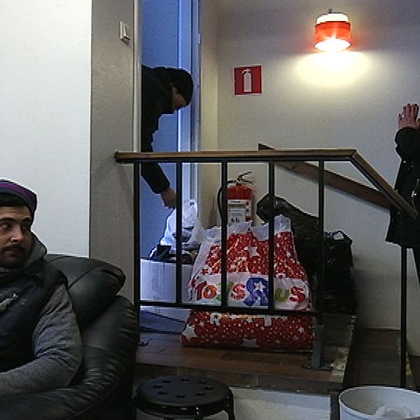 En man sitter i en soffa och håller i en kopp. En kvinna står vid en dörr i bakgrunden