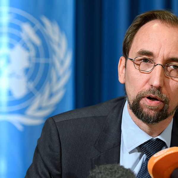 FN:S kommissionär för mänsliga rättigheter klädd i mörk kostym, sittandes framför mikrofoner med en blå bakgrund med FN-symbolen.