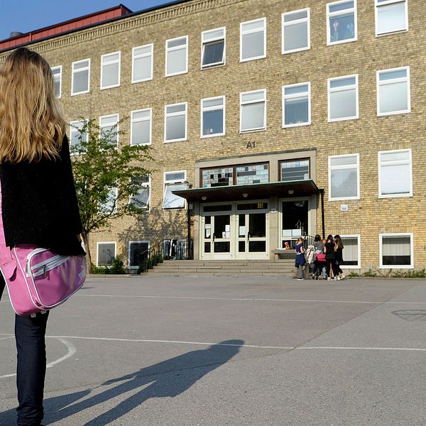 En ensam flicka på väg till skolan över en tom skolgård där en grupp flickor står vid ingången till skolbyggnaden