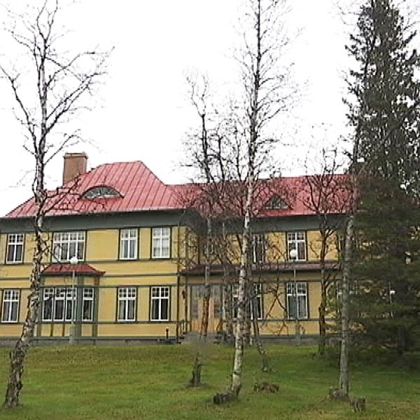Ett gult hus med massa fönster bakom träd och granar. En flaggstång vid sidan av huset.