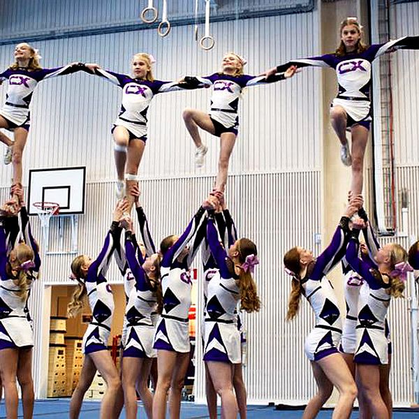 En stor grupp cheerleaders stor på golvet och håller upp sex andra som håller i varandras armar och tillsammans bildar en formation