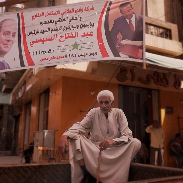 En man sitter utomhus på en mur i Kairo, nedanför en valbanderoll tillhörande presidenten al-Sisi.