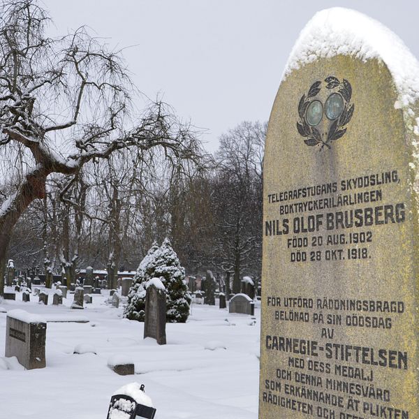 En gravsten med snö på toppen. I bakgrunden syns ett mörkt träd och en kyrkogård. På gravstenen kan man se en medalj, både fram- och baksida, i en inhuggen lagerkrans. På stenen står det: ”Telegrafstugans skyddsling, boktryckerilärlingen Nils Olof Brusberg. Född 20 aug 1902. Död 28 okt 1918.”