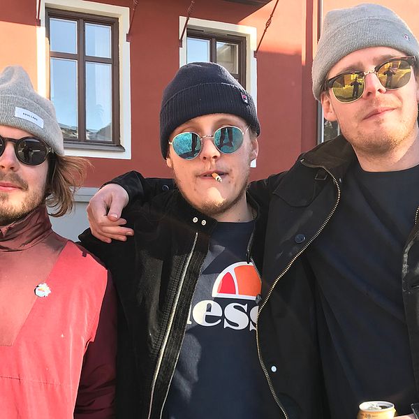 Tre killar med solglasögon, en med en cigarett i munnen och en med en ölburk i handen, står med armarna om varandras axlar.