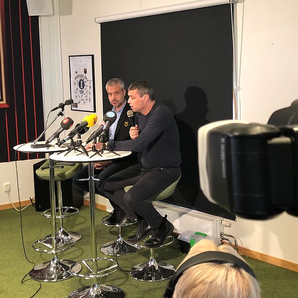 två män sitter vid mikrofoner i ÖFK-lokal, fotografer syns på sidan