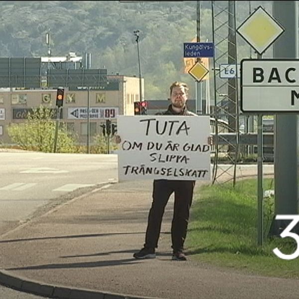 Reportern står i vägkorsning bredvid vägskylt ”Backadalsmotet” med handskrivet plakat: ”Tuta om du är glad att slippa trängselskatt”