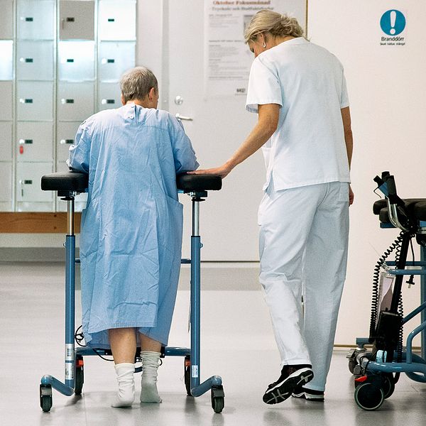 En patient med rullator går i sjukhuskorridoren tillsammans med personal.