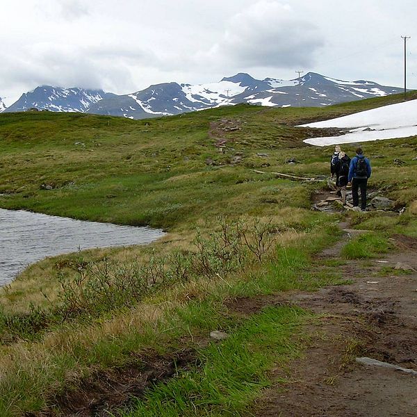 två personer vandrar på en led i fjällmiljö, förbi en sjö, en snöfläck. berg i bakgrunden