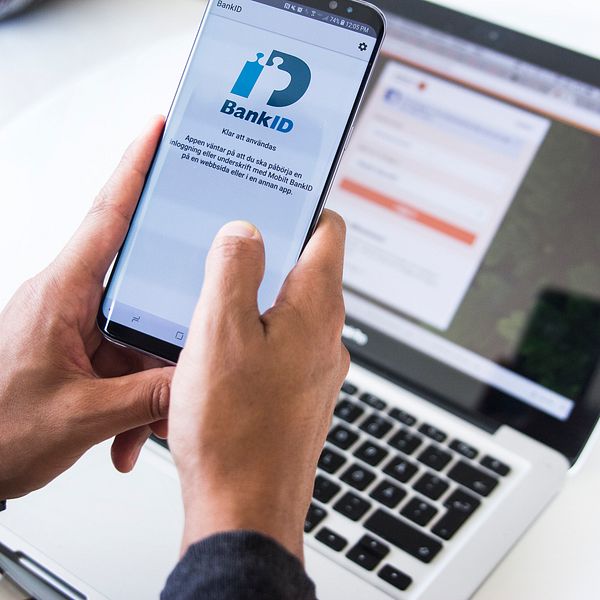 Två händer håller i en mobil framför en dator. På mobilskärmen syns mobilt bank-id:s logga.