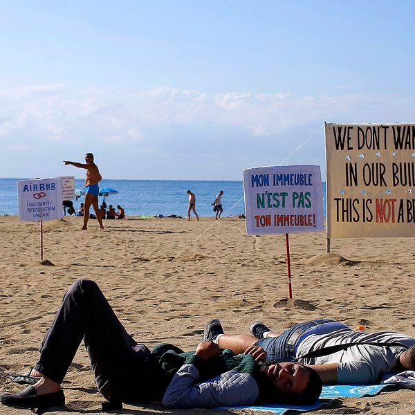 Två män ligger på en spansk sandstrand omgivna av skyltar som protesterar mot att turister bor i vanliga bostadshus.