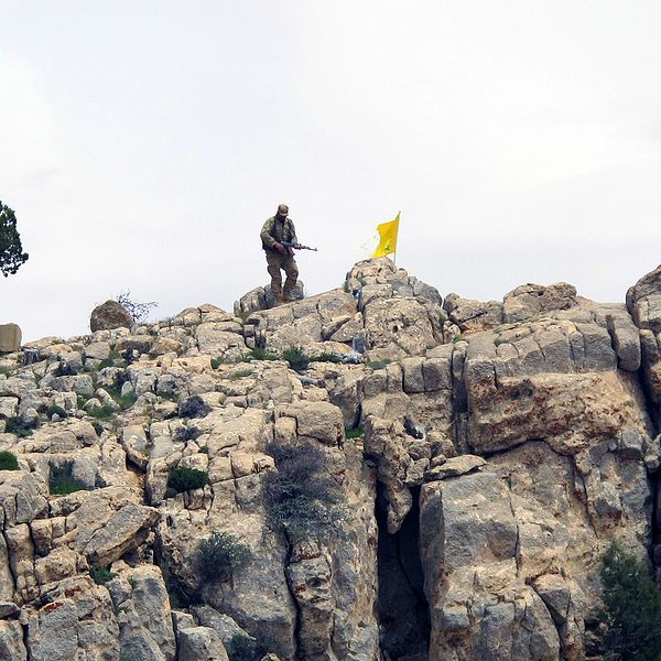 En hizbollahsoldat står på ett berg i Syrien, bredvid honom vajar organisationens flagga.