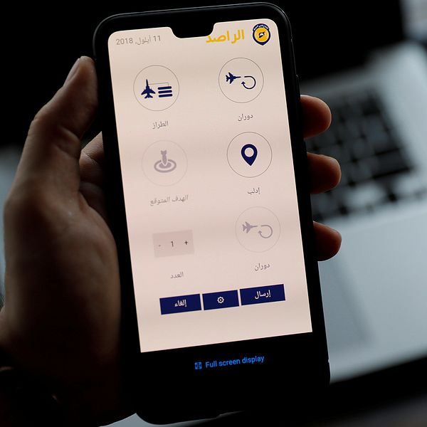 En hand håller upp en mobil, på skärmen syns appen som varnar för flyganfall i Syrien.