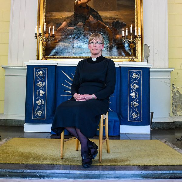 kvinna i prästkläder sitter på stol framför altaret i kyrka