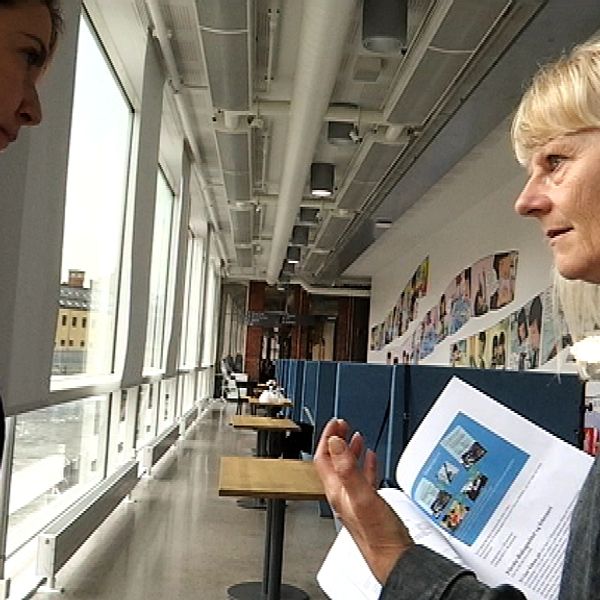 Två personer står och diskuterar, med papper i handen. I bakgrunden en korridor på högskolan.