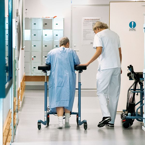 En kvinnlig patient med en rollator får hjälp av personal att ta sig fram i en korridor på ett sjukhus