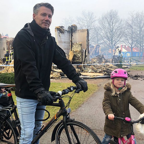 En pappa med två barn i förskoleåldern står med en cykel, i bakgrunden syns förskolan som nästan brunnit ner till grunden, det är brandrök i luften.
