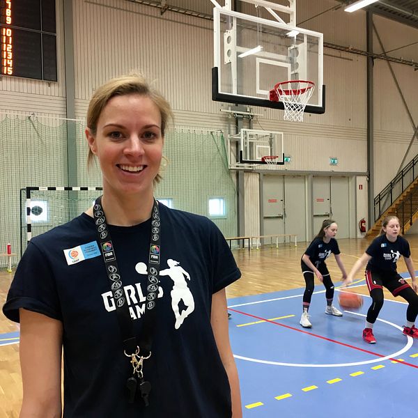 Elisabeth Egnell har en t-shirt som det står ”girlscamp” på och ler mot kameran. I bakgrunden tränar några av deltagarna i girlscamp.