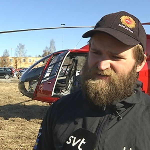 en man i keps och jacka står framför en helikopter på gräsyta, bebyggelse i bakgrunden