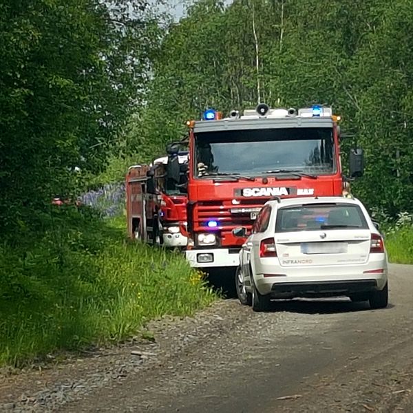 Två brandbilar syns på en grusväg med lövskog på båda sidor. En vit bil står parkerad framför räddningsfordonen.