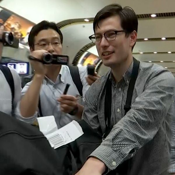 29-årige Alek Sigley anländer till flygplatsen i Peking i Kina omringad av journalister. Bilden är tagen ur en video.