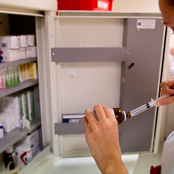 Närbild på tjej i profil med vit vårdrock som förbereder en spruta med medicin. Hon står framför ett medicinskåp.